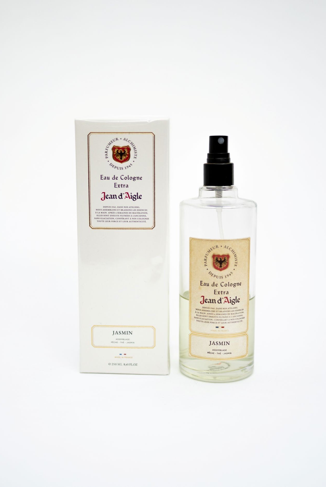 A clear glass perfume bottle with a black spray top stands next to its white rectangular box. Both feature cream labels with a crest and the text 'Jean d'Aigle' and 'Jasmin'.