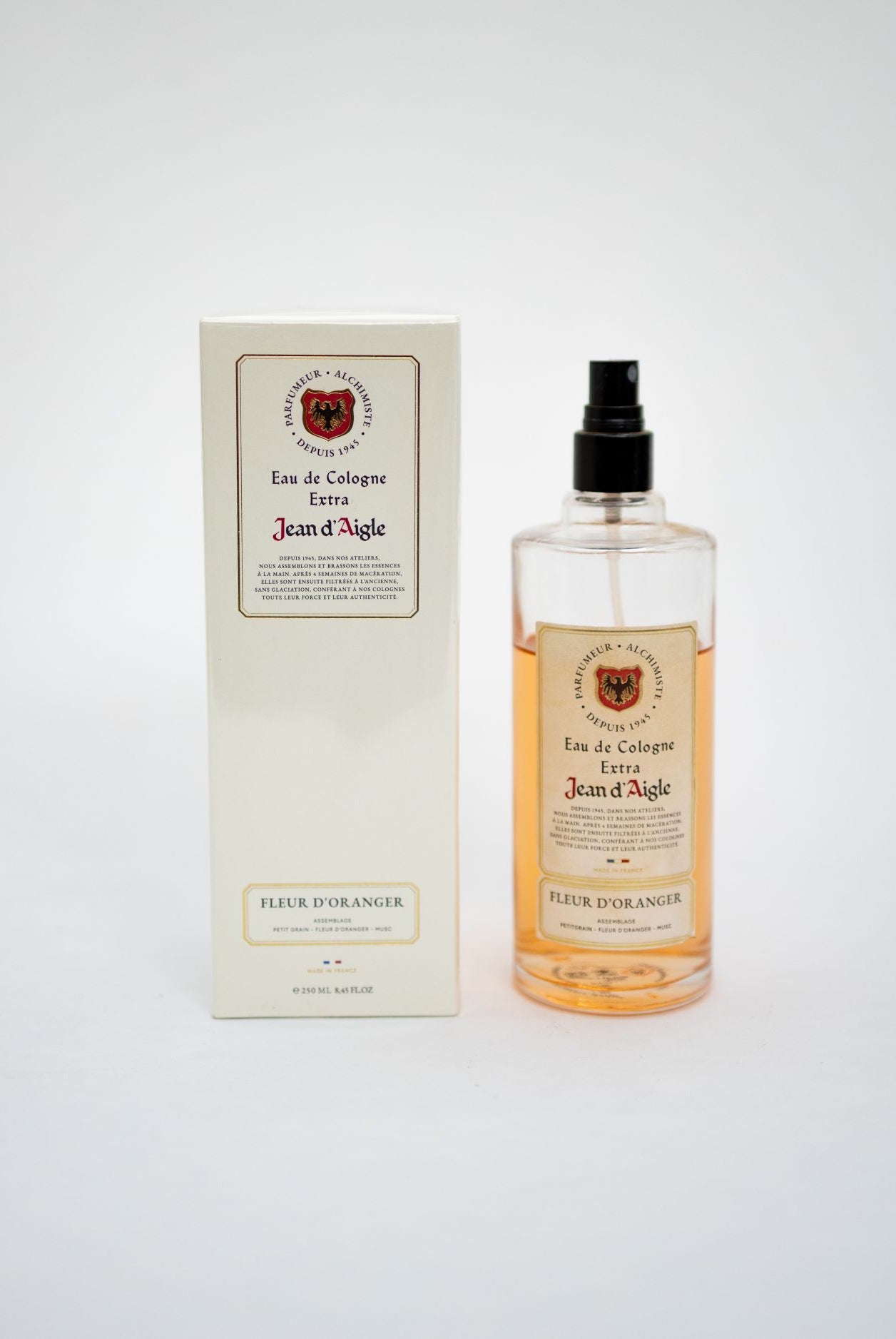 A bottle of Jean d'Agache Eau de Cologne Extra with amber liquid and a black spray top, standing next to its cream-colored packaging box on a light gray background.