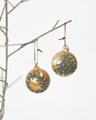Two round gold Christmas ornaments with hand-painted green pine tree and wreath designs, hanging from a thin decorative branch against a white background.