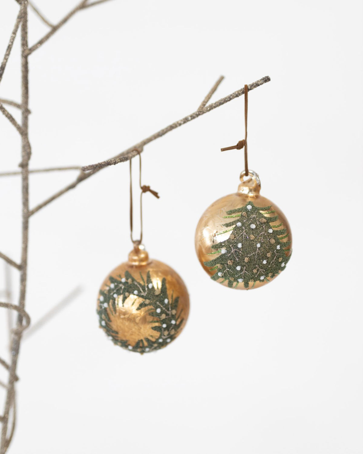 Two round gold Christmas ornaments with hand-painted green pine tree and wreath designs, hanging from a thin decorative branch against a white background.
