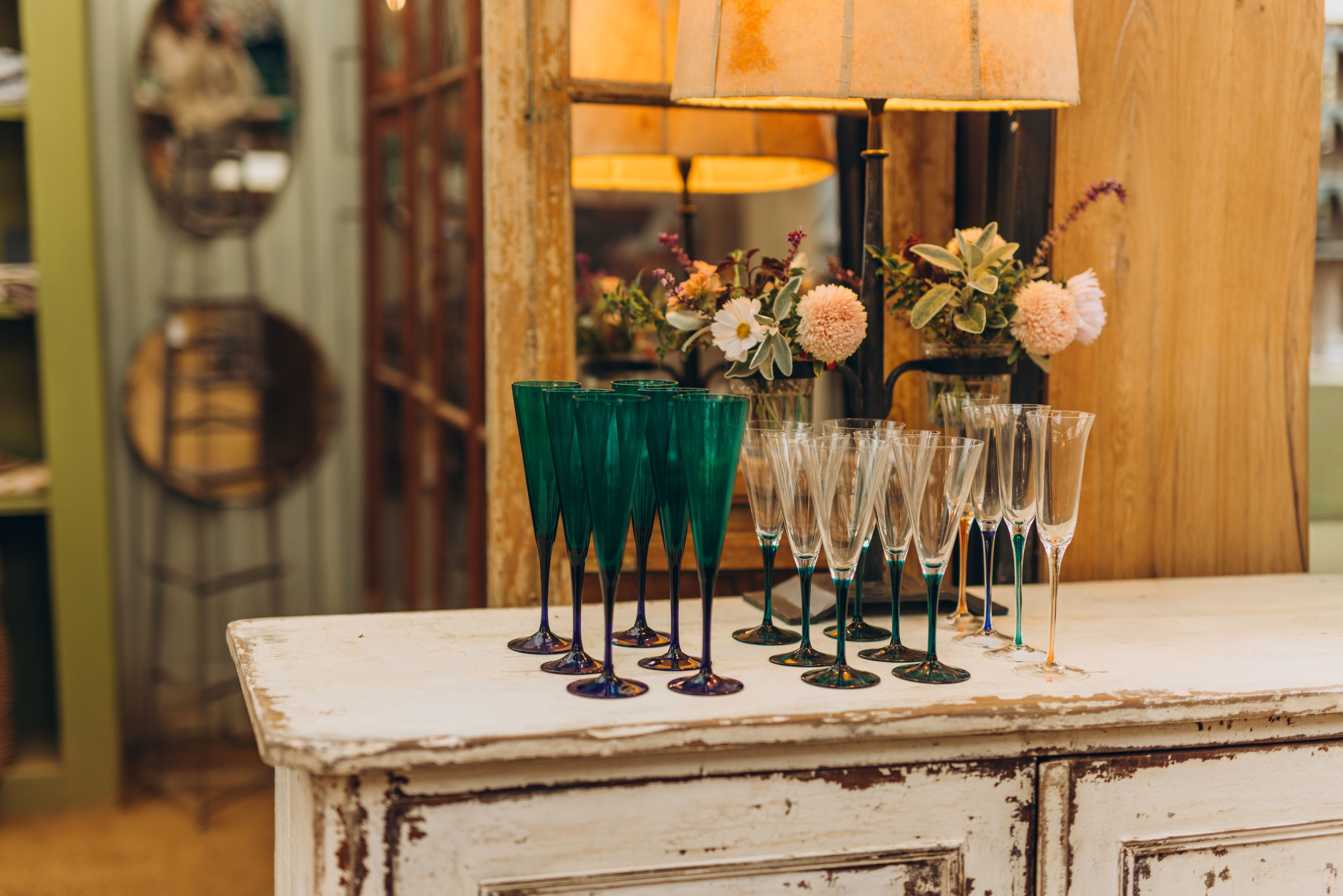 A set of champagne flutes and green goblets arranged on a distressed white wooden cabinet in a warmly lit, rustic interior with a lamp and flowers in the background.