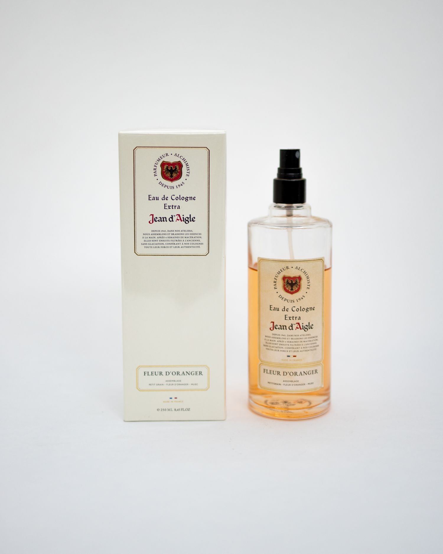 A bottle of Jean d'Agache Eau de Cologne Extra with amber liquid and a black spray top, standing next to its cream-colored packaging box on a light gray background.